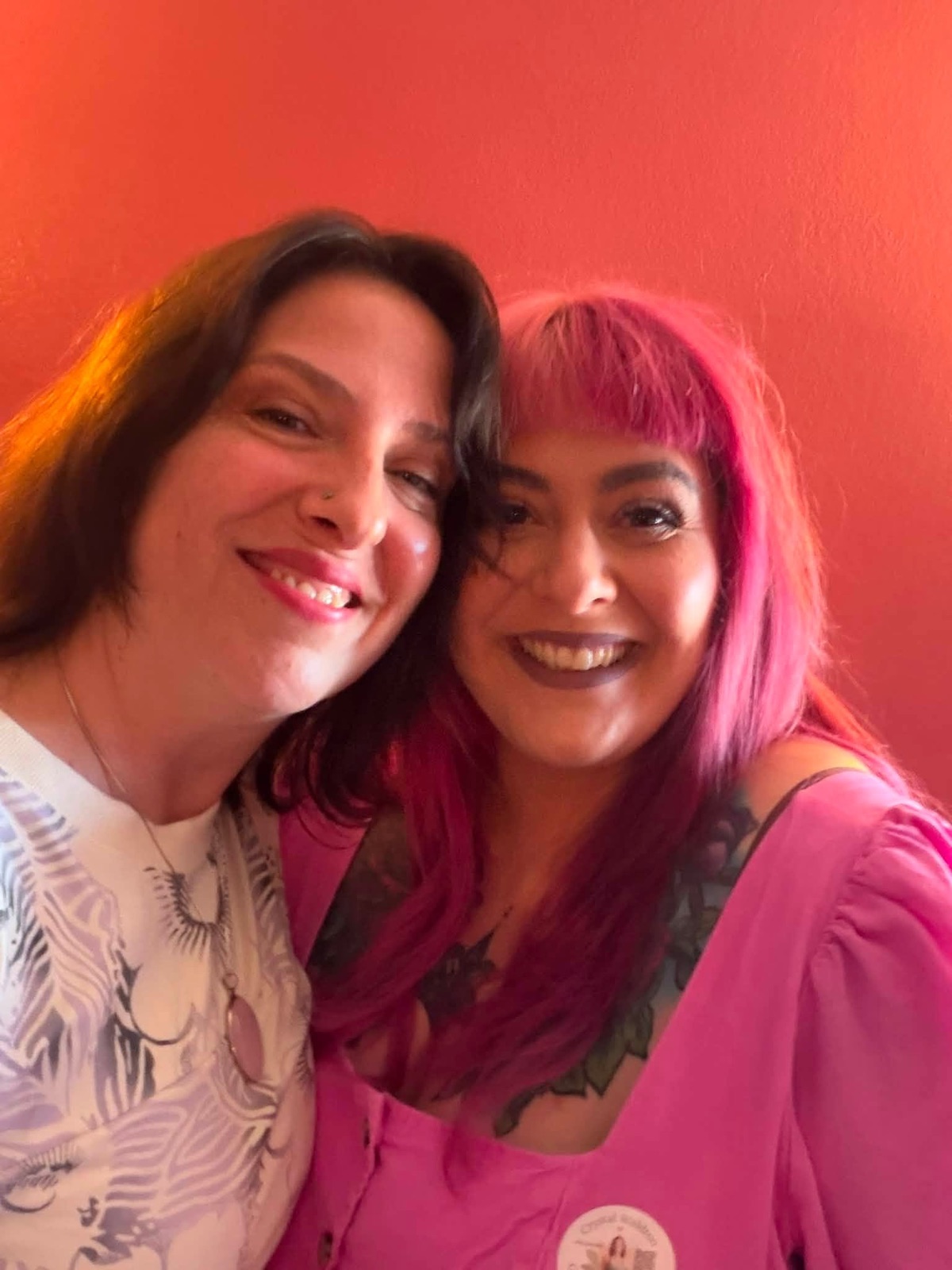 Two friends smiling together, one wearing bright pink, inside the studio.